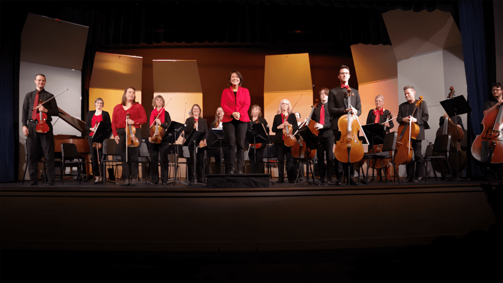 A group of musicians on stage, each holding various string instruments including violins, cellos, and double basses. They are dressed in black and red and appear to be finishing a performance or rehearsal. The background features lit panels in yellow and orange hues.