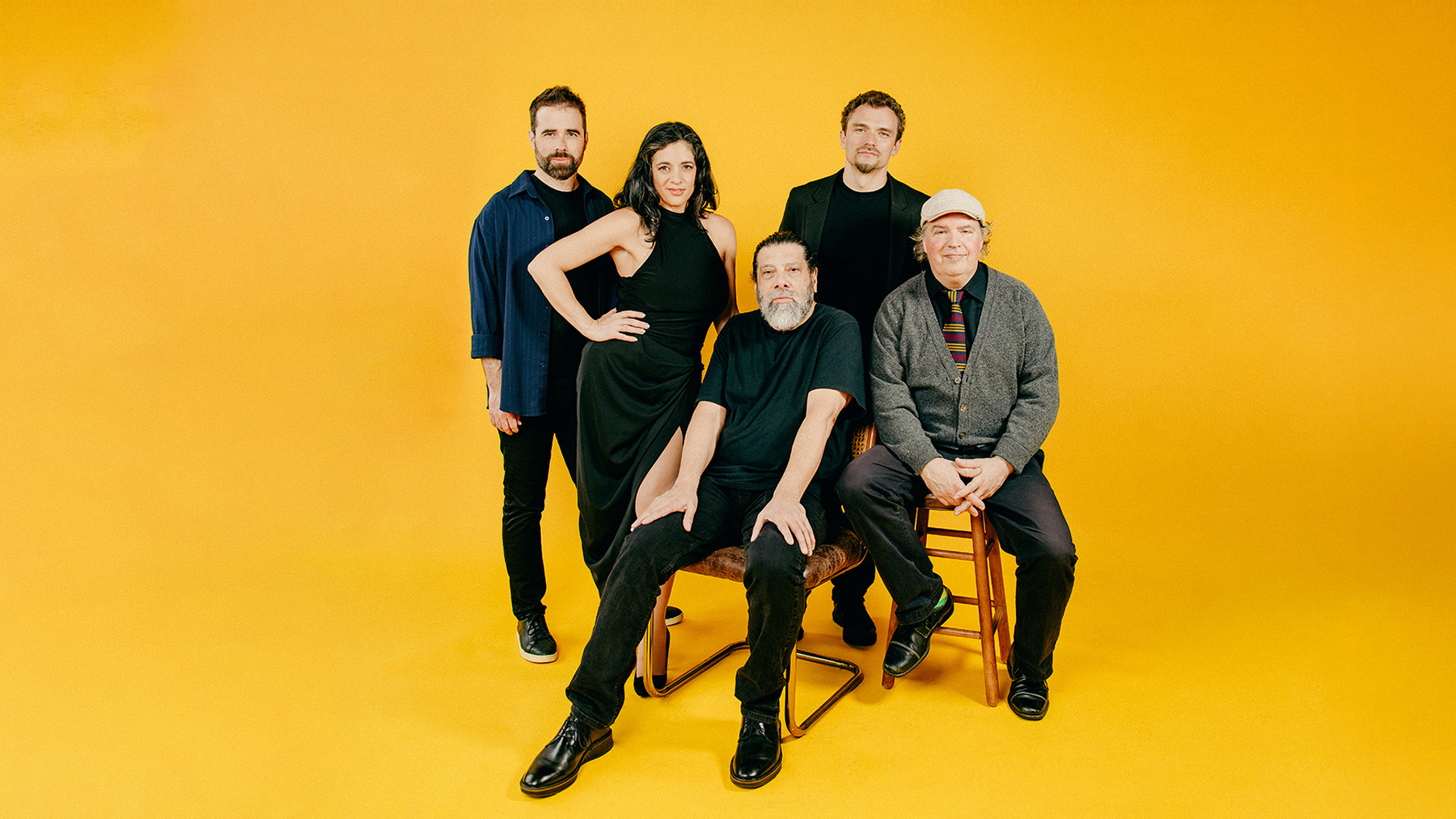 Five adults posing against a bright yellow background; three men are standing, one woman stands with a hand on her hip, and two men sit on chairs in front. All are dressed in dark or neutral clothing.