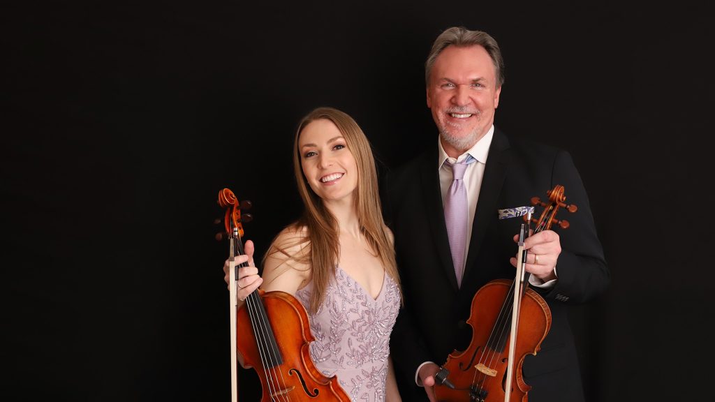 A smiling woman in a light purple dress and a smiling man in a suit hold violins against a plain black background.