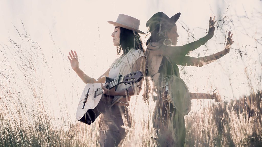 Two women stand back to back in a sunlit field, one holding a guitar. The photo is artistically double-exposed, creating layered images of both women with their arms raised, blending with tall grass.