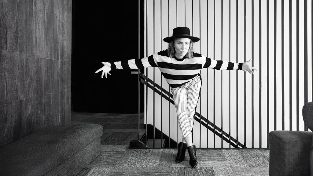 A person in a striped sweater, striped pants, and a wide-brimmed hat poses playfully with arms outstretched in a modern room with vertical lines on one wall. The image is in black and white.