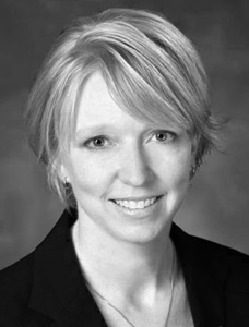 A professional headshot of a woman with short, light-colored hair, wearing a dark blazer and smiling at the camera. The photo is in black and white with a neutral background.