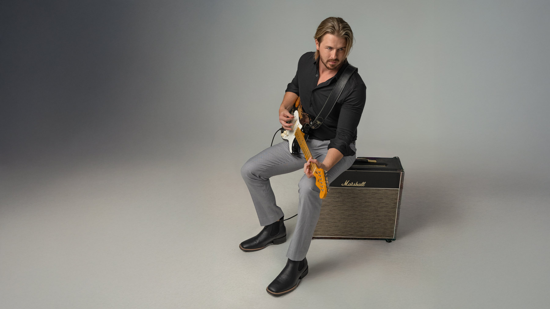 A man with blond hair and a beard, dressed in a black shirt and gray pants, sits on a Marshall amplifier and plays an electric guitar. The background is plain and softly lit.