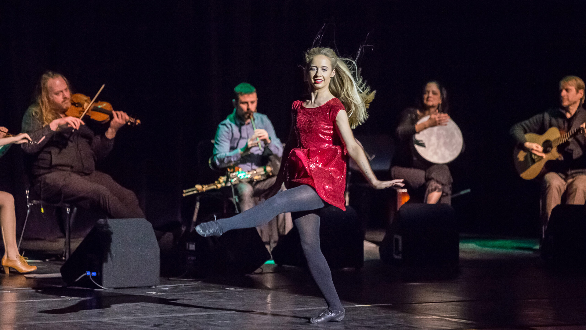 A woman in a red dress and black tights performs a lively dance on stage, kicking her leg high. Four musicians seated behind her play instruments including a violin, flute, drum, and guitar under stage lighting.