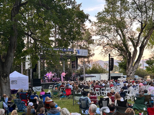 A large crowd sits on lawn chairs and blankets in a park, watching a live outdoor concert on a stage surrounded by trees at sunset. The atmosphere is lively and relaxed.