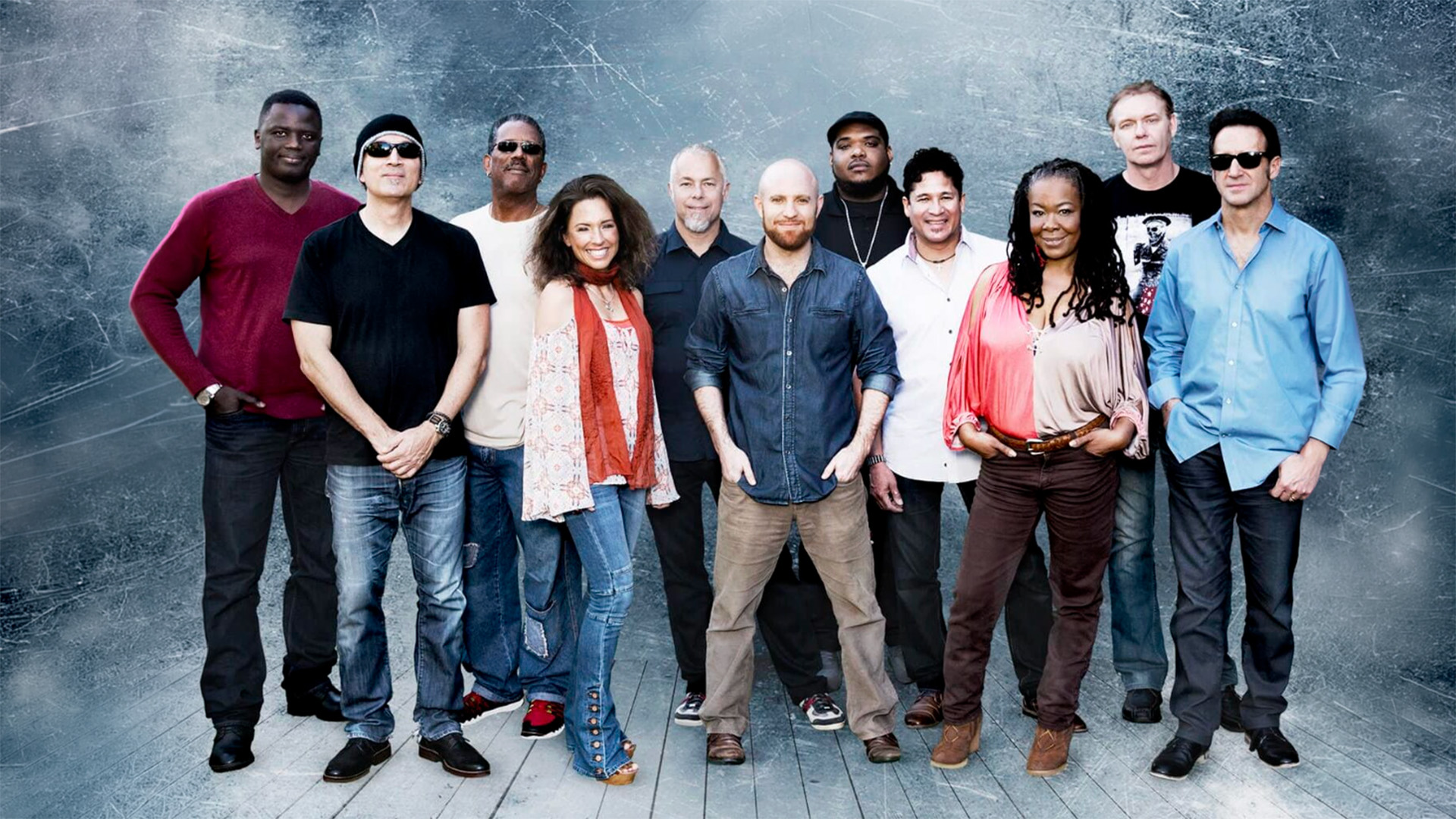 A group of eleven adults, consisting of men and women, stand together smiling in front of a textured blue background on a wooden floor. They wear casual clothing and appear relaxed and friendly.
