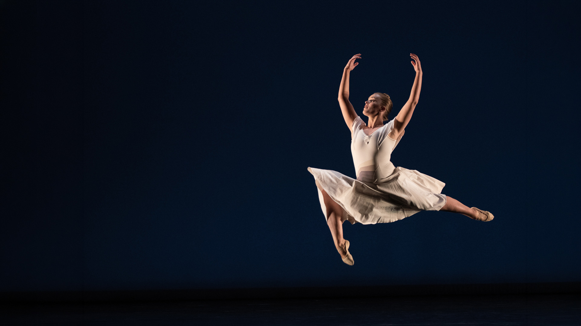 A ballet dancer in a white leotard and skirt leaps gracefully through the air on a dark stage, arms raised and legs extended, illuminated by stage lighting.