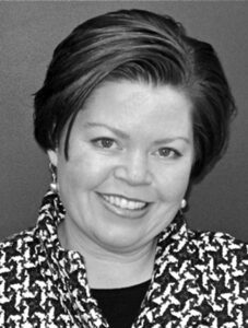 A woman with short, dark hair smiles at the camera. She is wearing a patterned jacket with a houndstooth design and pearl earrings, and is posed in front of a plain, dark background.