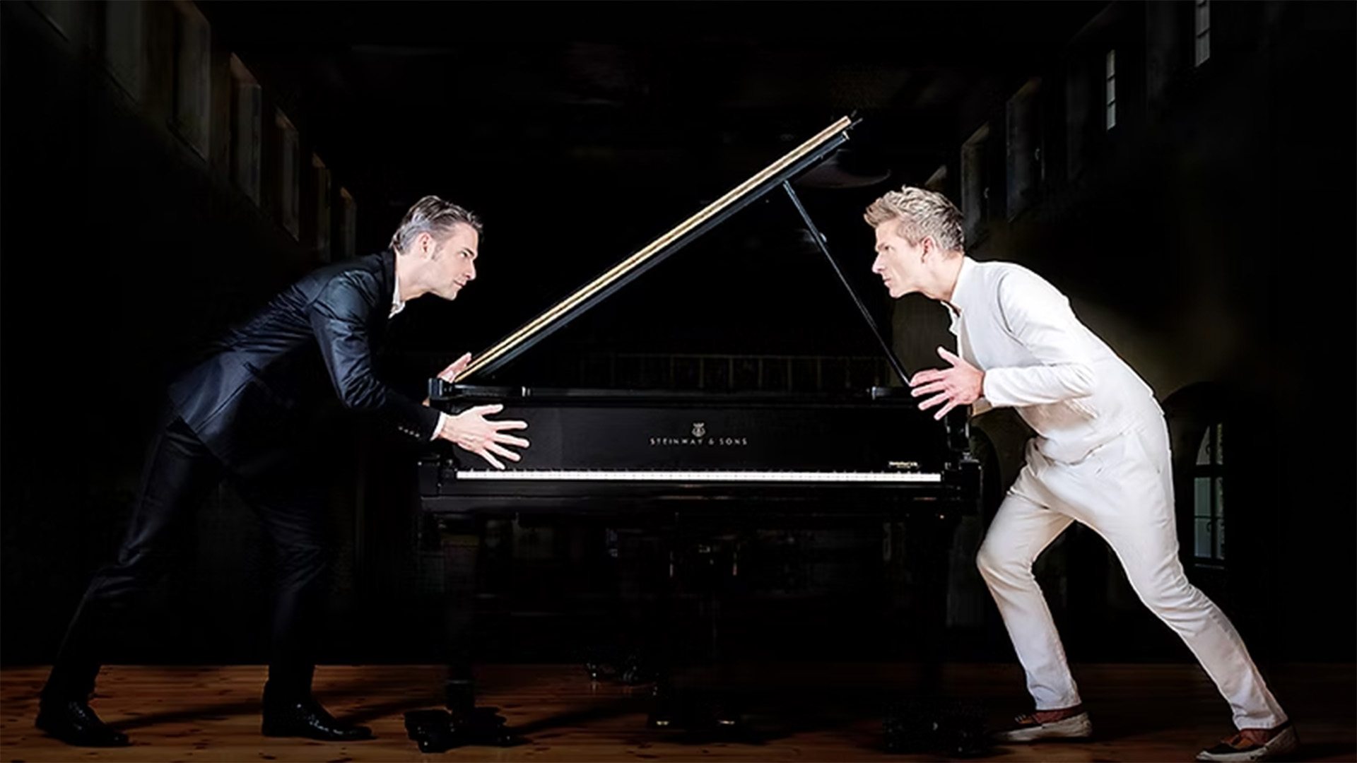 Two men, one in a black suit and one in a white suit, face each other intensely across a grand piano, each with hands on the piano as if preparing for a musical duel.