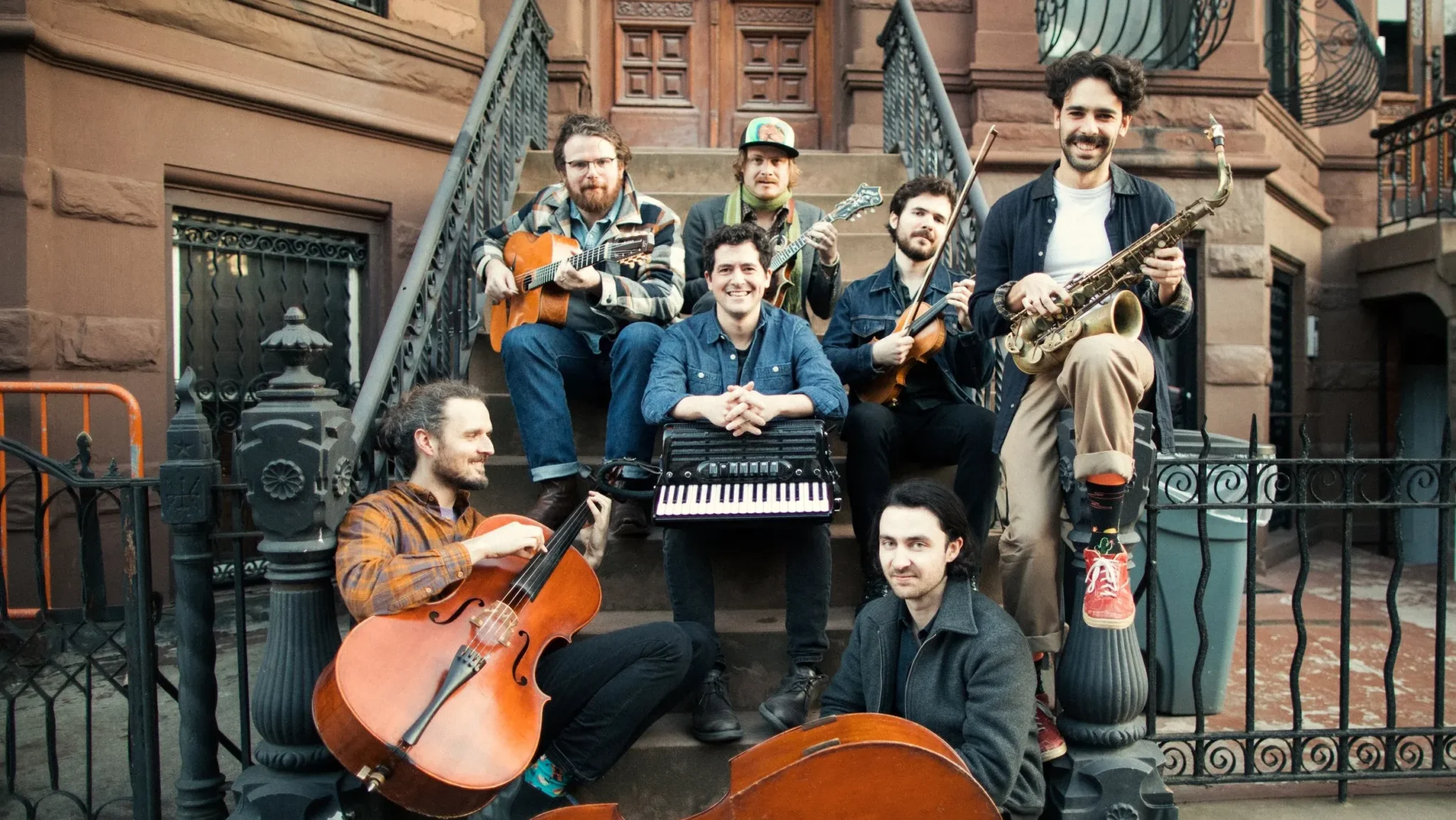 Seven musicians pose on brownstone steps with their instruments, including a cello, saxophone, keyboard, guitars, and mandolin. They smile at the camera in a relaxed, urban outdoor setting.