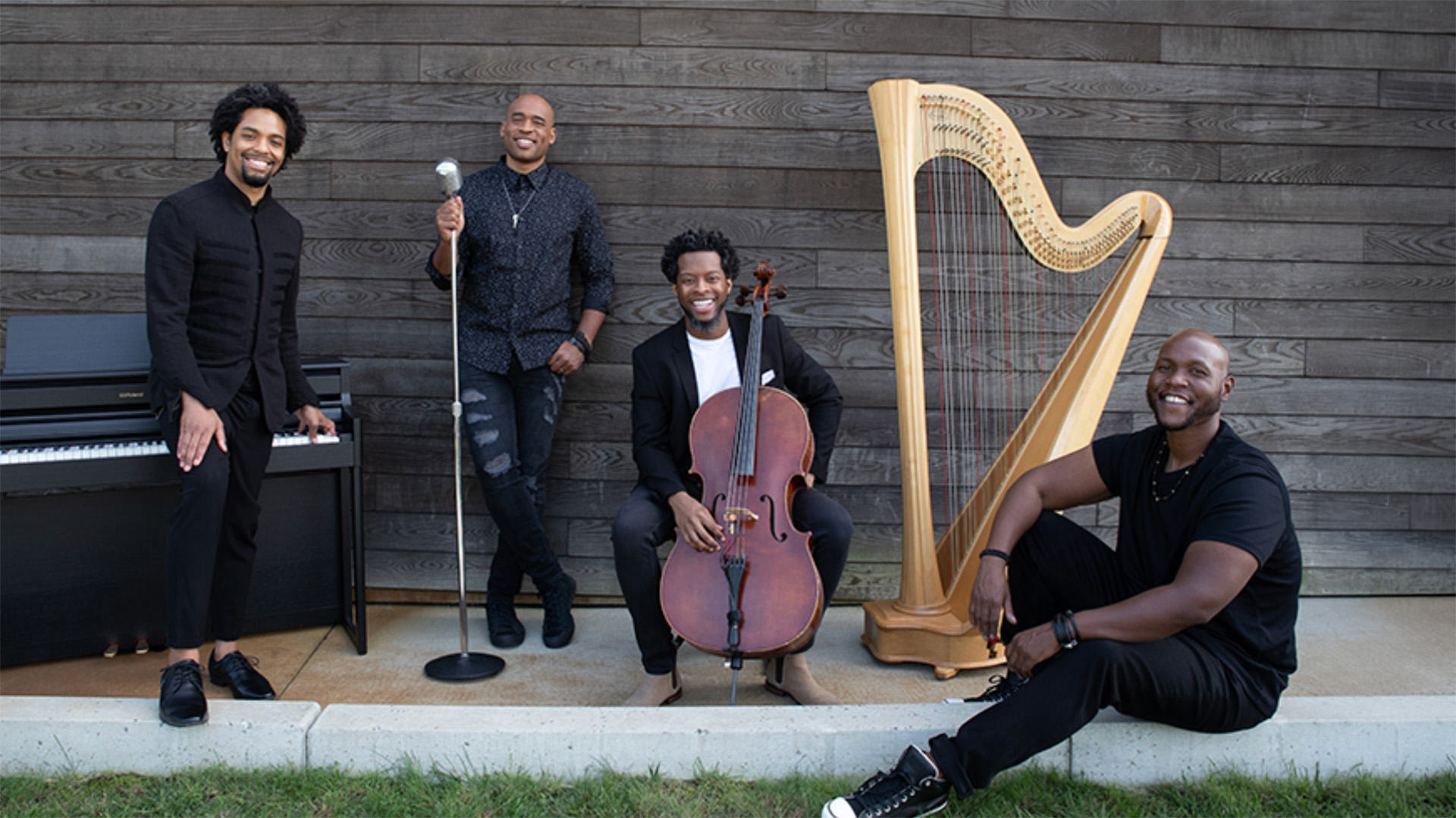 Four men pose smiling outdoors in front of a wooden wall with musical instruments: a keyboard, microphone, cello, and harp. The group appears relaxed and friendly, with some standing and others sitting on a low curb.