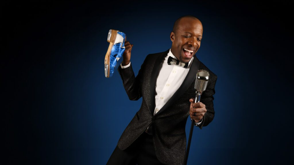 A man in a black tuxedo holds a blue tap shoe in one hand and sings passionately into an old-fashioned microphone, set against a dark blue background.
