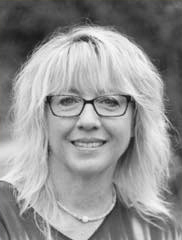 A woman with light, wavy hair and glasses smiles at the camera. She is wearing a necklace and a light top. The background is blurred and appears to be outdoors. The image is in black and white.