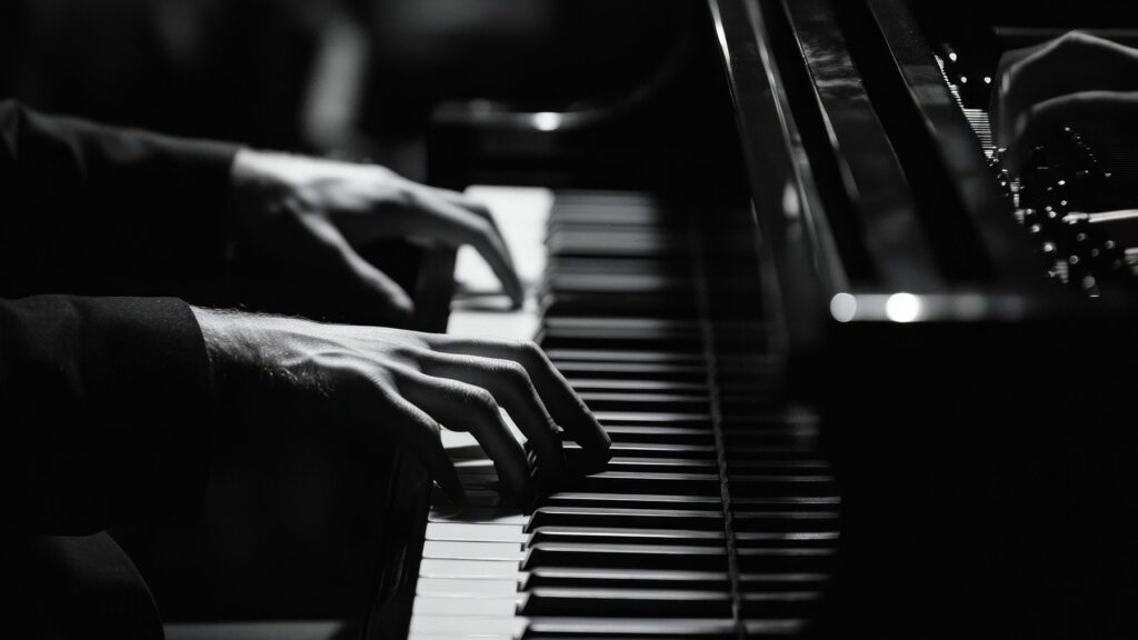 Close-up in black and white of a person’s hands playing a piano, with fingers pressing the keys and another pair of hands partially visible in the background.