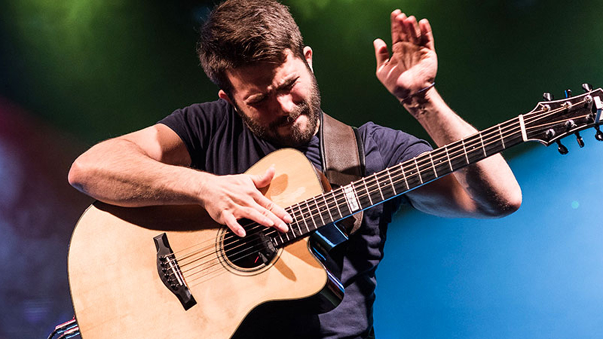 A man passionately plays an acoustic guitar on stage, using both hands to tap the strings, with eyes closed and a focused expression, under colorful stage lighting.