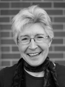 An older woman with short gray hair, wearing glasses and a scarf, smiles at the camera. The background is a brick wall. The image is in black and white.