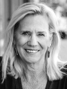 A smiling older woman with light-colored, shoulder-length hair wearing earrings, photographed outdoors in natural light. The image is in black and white.