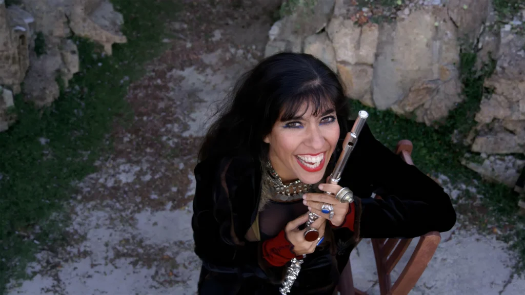 A woman with long dark hair smiles brightly at the camera while holding a silver flute. She is wearing dark clothing and jewelry, and is seated outdoors near rocks and grass.